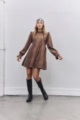 Vestido corto de gamuza color marrón, con diseño de corte evasé, volados en los hombros y mangas largas con puños elásticos fruncidos. Presenta un cuello alto y una falda con un volado en el ruedo.