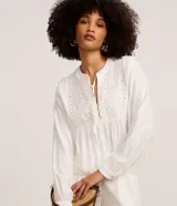 Blusa bata blanca de manga larga con cuello padre, lazo ajustable en el escote y botones frontales parciales. Presenta detalles de bordado floral y encaje broderie en la parte frontal del busto.