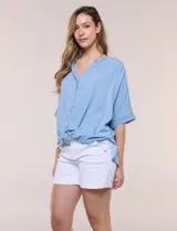 Blusa estilo denim de lyocell celeste, de silueta amplia y suelta, con escote en V, cartera frontal de botones y mangas cortas japonesas de hombros caídos. Cuenta con ruedo asimétrico y posibilidad de anudar al frente.