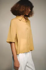 Camisa de color mostaza, con cuello solapa, manga corta y bajo recto. Cierre frontal con botones.