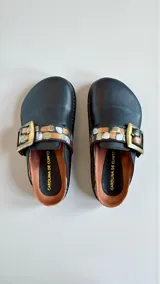Sandalias de cuero color negro, estilo zueco, con tira ajustable mediante hebilla metálica grande y detalles pintados a mano en tonos dorados, plateados y cobre.