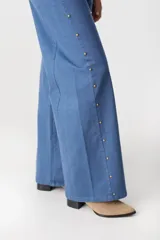 Jean de corte recto y tiro medio, color azul, con detalles de tachas metálicas doradas a lo largo de las costuras laterales externas.
