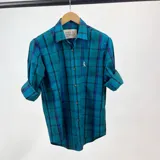 Camisa de manga larga a cuadros en tonos azul, celeste y negro. Tiene cuello clásico, cierre con botones y un pequeño logo bordado en el pecho. Las mangas pueden usarse arremangadas.