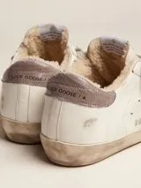 Championes Golden Goose modelo Super-Star confeccionados en cuero blanco con efecto desgastado. Destacan por su estrella lateral en glitter plateado, refuerzo del talón en cuero marrón y forro interior de lana tipo shearling. Incluyen cordones grises y suela de goma con acabado envejecido.