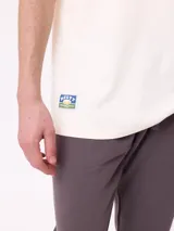 Remera de manga corta color crema con estampado gráfico en la espalda que representa un paisaje de montaña con un lago, árboles y una carpa, junto con el logo de la marca Rusty.