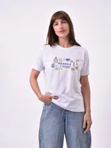 Remera blanca de manga corta con cuello redondo y un estampado central que incluye la frase "ISOLA BELLA" rodeada de ilustraciones de utensilios de cocina y cerámica en tonos azules y terracota.