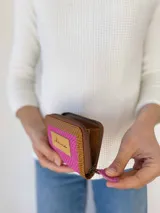 Billetera compacta de cuero con diseño bicolor en fucsia y marrón, con textura grabada tipo reptil. Cuenta con cierre metálico perimetral, tirador de cuero fucsia y etiqueta frontal con logo grabado.