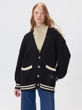 Cardigan de punto trenzado en color negro con detalles en color crema en los bolsillos, puños y ruedo. Presenta un diseño oversize, cierre frontal con botones y bordado decorativo en uno de los bolsillos delanteros.