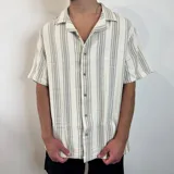 Camisa de manga corta con corte oversize, cuello tipo cubano y cierre frontal con botones. Presenta un diseño de rayas verticales en tonos crudo y gris.