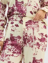 Conjunto de dos piezas compuesto por camisa de manga larga con cuello solapa y botones frontales, y pantalón de tiro alto con pierna recta y bolsillos laterales. Ambas prendas presentan un estampado paisajístico en tonos marrones sobre fondo crema.