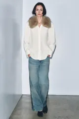 Camisa de popelín color crudo con cuello solapa y aplicación desmontable de pelo sintético en tono marrón. Presenta cierre frontal con botones, manga larga con puños y bajo asimétrico.