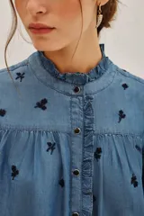 Camisa de lyocell color azul con estampado floral pequeño en tono oscuro. Presenta cuello alto con volados, cierre frontal con botones y mangas largas abullonadas con puños abotonados.