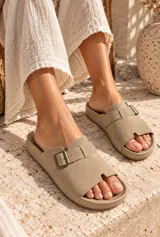 Sandalias planas color beige con tira ancha en el empeine, hebilla metálica decorativa y diseño abierto.