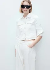 Chaqueta crop de denim blanco, confeccionada en algodón con un toque de elastano. Presenta cuello de solapa, cierre frontal con botones metálicos, bolsillos de parche con solapa en el pecho y manga larga con puños abotonados.