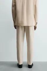 Pantalón chino de corte relajado, color beige claro, con textura de rayas verticales sutiles. Presenta cintura ajustable con cordón interior, bolsillos delanteros y bolsillos traseros de vivo.