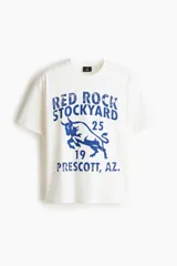 Remera de algodón blanca con estampado azul estilo universitario de toro y texto "Red Rock Stockyard". Presenta cuello redondo, mangas cortas y hombros caídos.