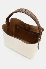 Bolso tipo saca color crudo con cuerpo en tejido canvas, asa corta de mano trenzada en marrón y correa bandolera adicional. Cuenta con cierre de botón imantado y bolsillo interior con cremallera.