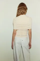 Top entallado de punto suave en color beige, con cuello tortuga, capa decorativa superpuesta y frunces laterales que generan un efecto drapeado.