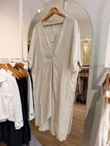 Vestido midi de lino color beige, con diseño oversize, escote en V y detalle de nudo drapeado en el frente. Posee mangas cortas amplias con dobladillo y corte asimétrico, siendo más largo en la parte trasera.