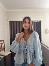 Camisa de tela liviana color celeste, con escote en V, mangas largas abullonadas con puños elásticos y detalle de volado fruncido en el frente con cierre de cordones ajustables.