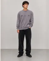 Buzo oversize de cuello redondo en color gris, con diseño estampado en el frente que incluye logotipos y texto. Presenta puños y ruedo acanalados, con una pequeña etiqueta decorativa en la parte inferior.