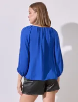 Blusa de gasa color azul, con cuello redondo y aberturas frontales en V. Presenta mangas raglán con apliques de puntilla a lo largo y puños fruncidos.