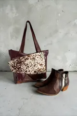 Cartera de cuero marrón estilo tote bag, con diseño en patchwork y bolsillo frontal de pelo con estampado animal print tipo vaca. Cuenta con doble asa de hombro y detalles metálicos.
