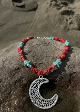 Collar corto de escallas de piedra coral en tonos rojizos, intercaladas con piedras turquesas y pequeños apliques plateados. Cuenta con un gran dije colgante de metal plateado con diseño de media luna texturada y calada.