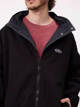 Campera reversible con capucha oculta, confeccionada en sarga de algodón con elastano en el exterior y nylon de poliéster en el interior. Presenta cierre frontal con botones a presión, costuras visibles en contraste y puños elásticos.