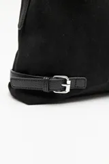 Cartera de gamuza negra con textura suave y acabado aterciopelado. Cuenta con una silueta amplia y flexible, cierre superior metálico, correa regulable y detalles laterales con hebillas.