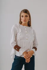 Camisa blanca de manga larga con detalles de alforzas y volados en el frente, cuello redondo fruncido con cierre frontal de botones y puños acampanados.