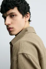 Camisa de manga larga color marrón verdoso, con cuello solapa y cierre frontal de botonadura. Presenta un tejido con textura rústica y un calce relajado.