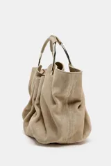 Bolso tipo saca de serraje color beige con doble asa rígida asimétrica y asa bandolera extraíble. Incluye cartera con bolsillo extraíble.