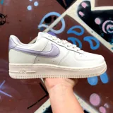 Championes Nike Air Force 1 de corte bajo, con cuerpo de cuero blanco, suela de goma color crema y detalles en tono lila metalizado en el swoosh lateral y el talón.