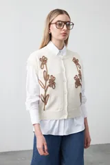 Chaleco tejido color crema con cuello redondo y cierre frontal de botones. Presenta un diseño bordado en relieve de color marrón con motivos florales y orgánicos en el frente.