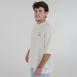 Remera color crema de corte holgado con cuello redondo y mangas cortas. Estampado pequeño en el pecho con texto en azul y amarillo.