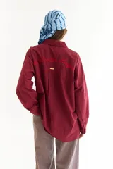 Camisa de corte oversize color bordó, con cuello camisero, mangas largas y bordado en hilo rojo en la espalda con la frase The Winter League. Presenta recortes con costuras a contratono y detalle de placa metálica pequeña en la parte superior trasera.