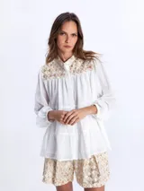 Camisa blanca vaporosa con cuello mao, botonadura frontal oculta y mangas largas abullonadas con puños elásticos. Presenta un diseño de paneles fruncidos y detalles bordados con motivos geométricos dorados en la parte superior delantera.