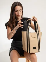 Bolso tipo tote de color beige con detalles en negro, confeccionado en material símil cuero con textura soft. Cuenta con doble asa de hombro, cierre con presilla e imán, y logo de la marca en la parte frontal. Incluye un necessaire interno con cierre.