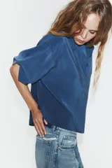 Camisa de corte fluido con cuello solapa y manga corta con dobladillo. Presenta un cierre frontal con botones ocultos bajo una tapeta y un diseño con bajo ligeramente más largo en la parte posterior.