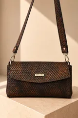 Cartera bandolera de cuero vacuno con textura tejida en tonos marrones y negros, solapa frontal con cierre magnético y placa metálica con la marca "Pecarí". Incluye correa larga de hombro desmontable con detalles de herrajes plateados.