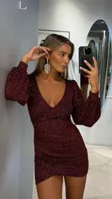 Vestido corto de fiesta confeccionado en tejido de lurex color bordó con destellos brillantes. Presenta un escote en V profundo, mangas largas abullonadas y un diseño ajustado al cuerpo con un corte cruzado en el ruedo.