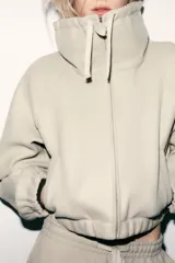 Sudadera tipo bomber de tejido suave, color beige, con cuello alto ajustable mediante cordones, cierre frontal con cremallera, mangas largas con puños elásticos y bolsillos laterales de vivo.