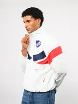 Buzo deportivo de nylon con medio cierre frontal y cuello alto. Presenta un diseño de bloques de color en azul, blanco y bordó, con el escudo del Club Nacional de Football y el logo de Umbro en el pecho.