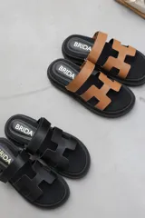 Sandalias planas de cuero sintético, con base negra y tiras anchas en forma de H en color caramelo o negro.