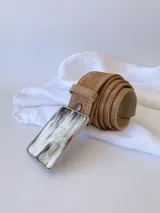 Cinto de cuero color marrón con forro de cuero y hebilla rectangular con diseño de guampa en blanco y negro con marco metálico plateado.