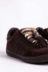 Zapatillas urbanas color tostado con textura geométrica en el cuerpo y detalles de gamuza en la puntera y laterales. Presentan un diseño distintivo con cordones anchos tejidos en tonos tostado y blanco, y un accesorio metálico con borla de gamuza en el empeine.