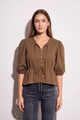 Blusa de algodón color marrón con diseño de broderie, escote en V con cordones ajustables, cierre frontal con botones y mangas tres cuartos abullonadas con puños elásticos. Presenta un corte ajustado en la cintura con cordón de ajuste.