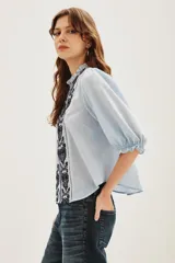 Camisa de algodón color celeste con cuello alto con volados, mangas tres cuartos abullonadas y cierre frontal con botones. Presenta un diseño con dos franjas verticales de bordado intrincado en color negro en el frente.
