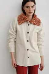 Camisa de paño color crudo, confeccionada en lana, con cuello tejido en tonos rojizos y naranjas, cierre frontal de botones y bolsillos frontales tipo parche.